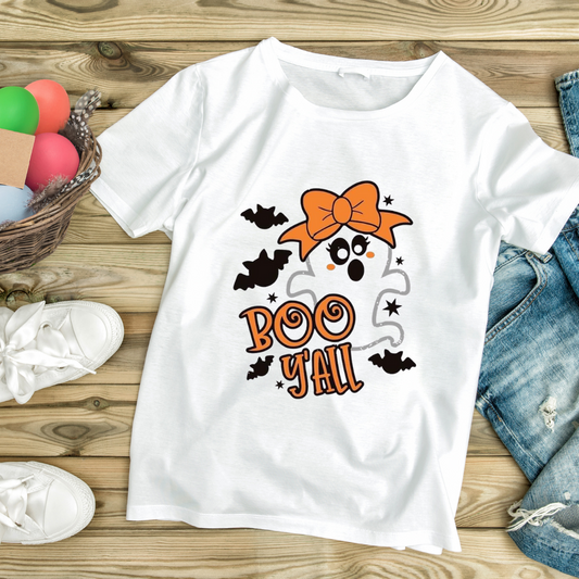 White funny ghost graphic tee with orange bow and Boo Y'all text laid on wooden surface with denim shorts and white sneakers